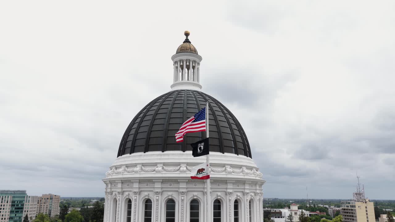 Californiens State Capitol-kuppel med vajende amerikansk flag i Sacramento, USA, med neoklassisk arkitektur, overskyet himmel og patriotisk atmosfære ved et statsligt vartegn. Kredsende luftfoto