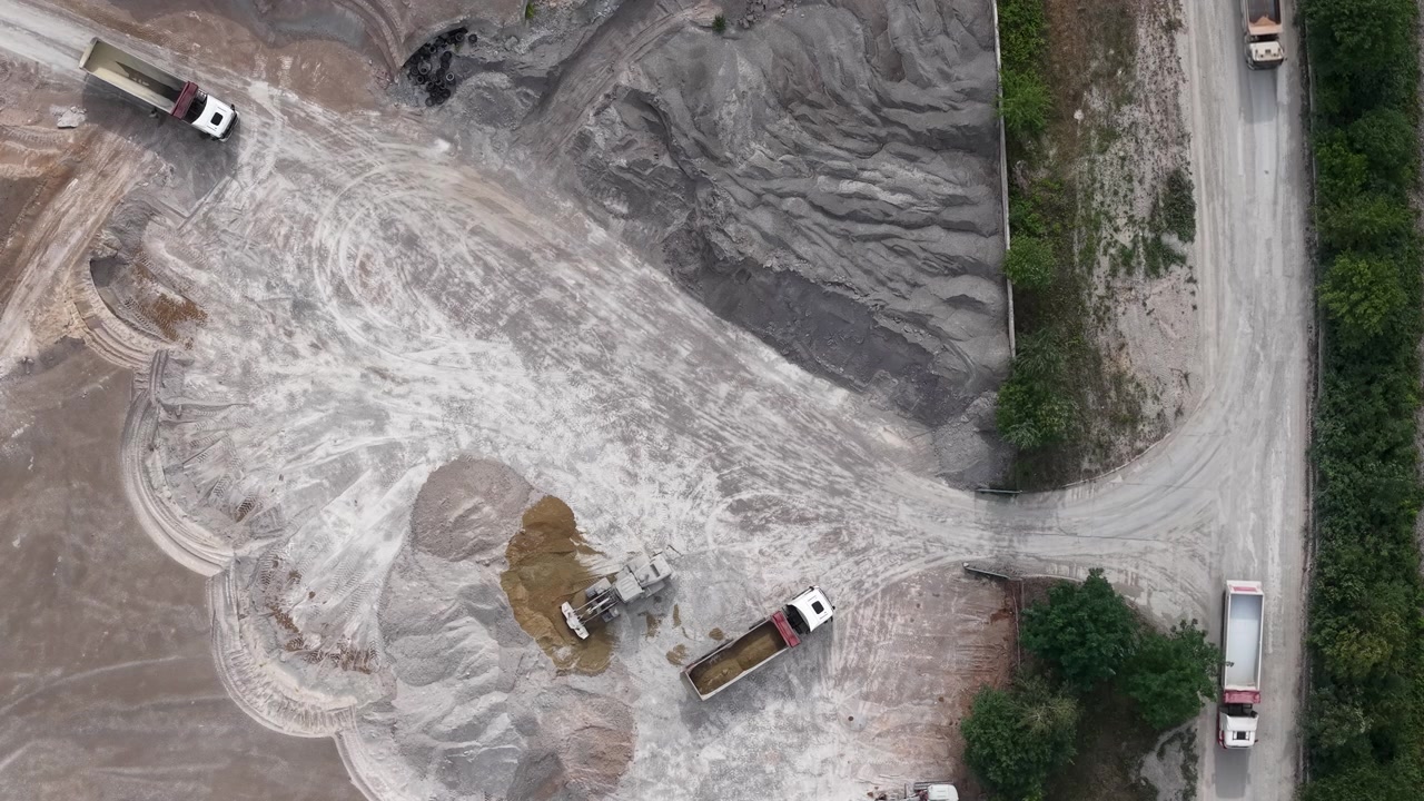 Aerial view of a quarry operation with trucks and an excavator moving materials on a dusty industrial site.
