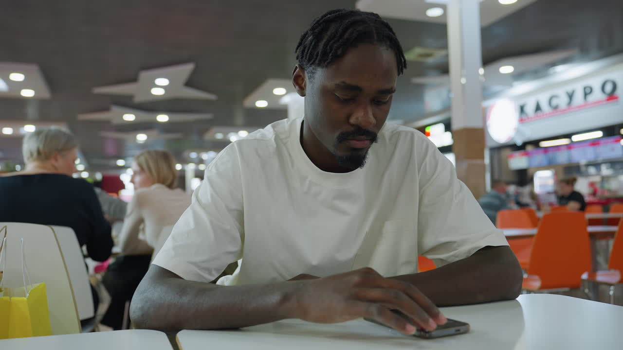 Young man with twisted hair sits alone at table using phone in crowded eatery, wearing white shirt, focused and thoughtful, with blurred background of chatting people, orange chairs, and lit signage
