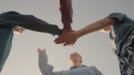 upward view of people in hoodies placing their hands together as team members with sunlight creating a glow effect around them, while they smile and embrace team spirit and unity