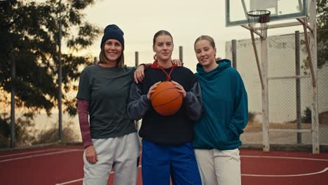 portrait of a trio of happy girls basketball players blondes in hoodies holding an orange basketball standing and posing on a red basketball court fenced with bars in the morning in the summer