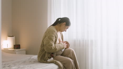 Loving-Young-Mother-Breastfeeding-Her-Newborn-Baby-While-Sitting-On-The-Bed-In-The-Morning-At-Home