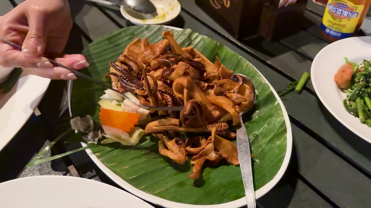 Hand serves deep fried squid with fork onto banana leaf platter at outdoor dining table