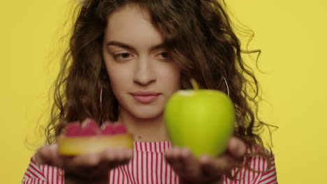 young woman weighing apple and cake in hands in yellow studio