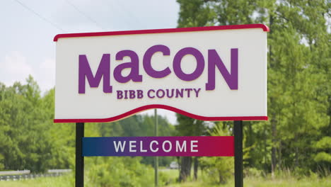 Macon-Bibb-County-Georgia-welcome-road-sign-in-front-of-highway-road-woods-telephoto-pan-up-left-truck-driving-behind-60p-0005