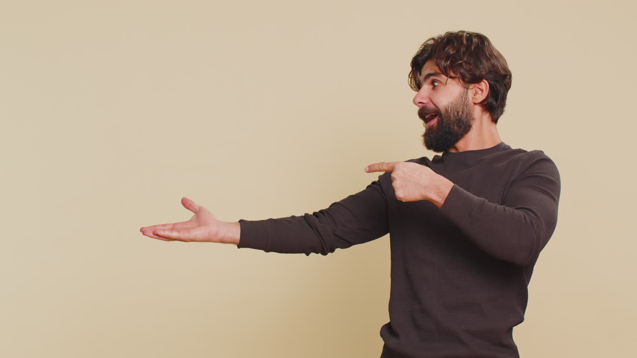 Lebanese young man showing thumbs up and pointing at empty blank place against beige background