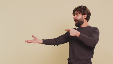 lebanese young man showing thumbs up and pointing at empty blank place against beige background