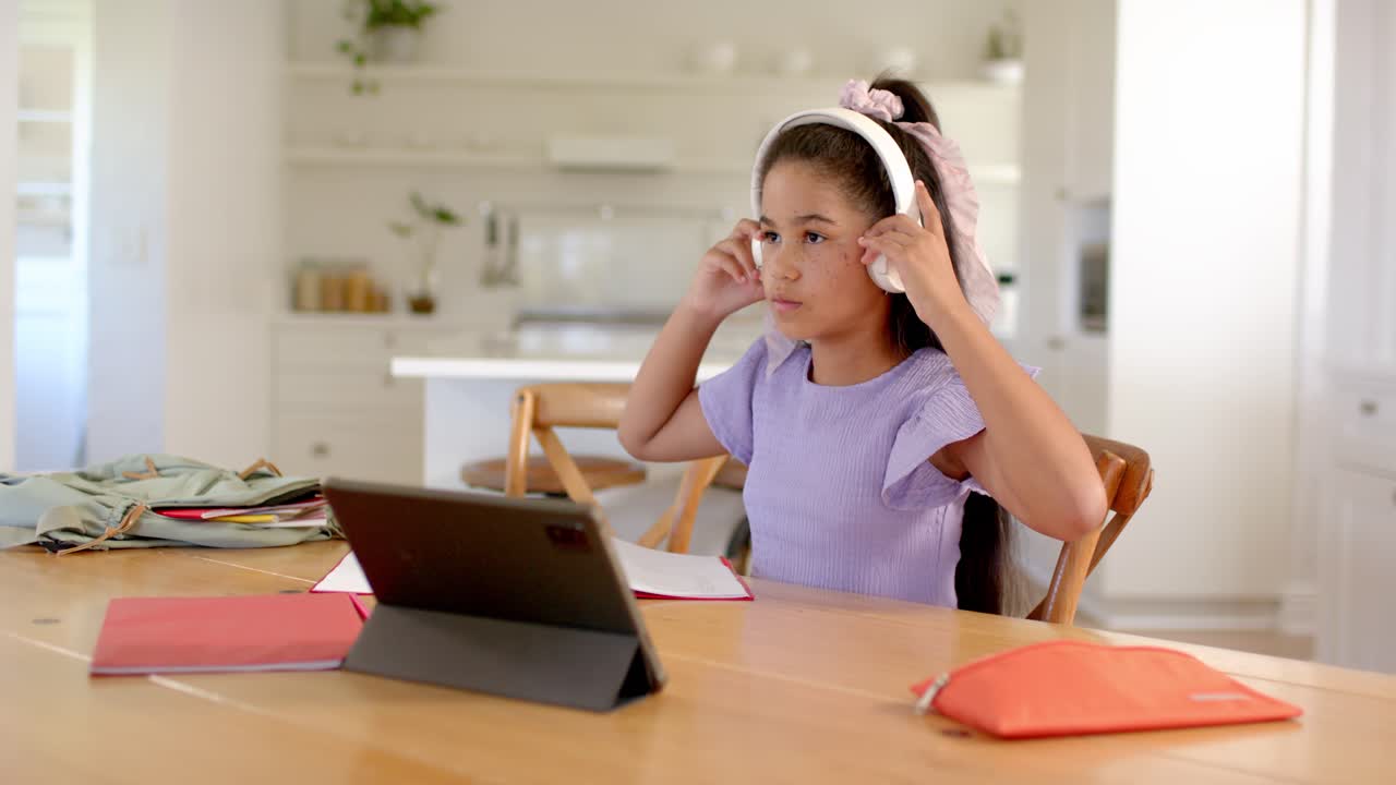 Putting on white headset, girl glancing at and steadying tablet for school in kitchen, purple top