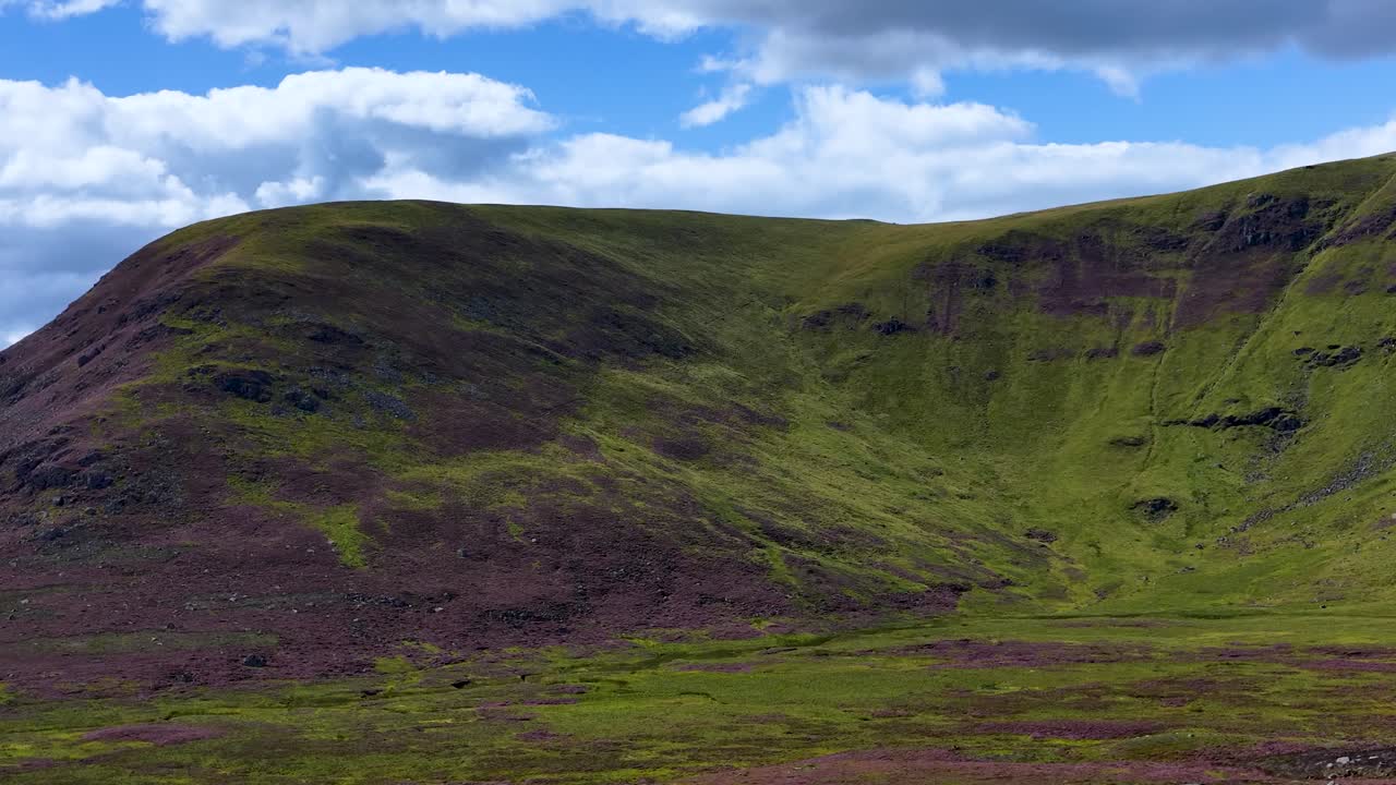Wide daylight pan over lush Scottish Highlands, heather fields, grassy slopes, and dramatic clouds