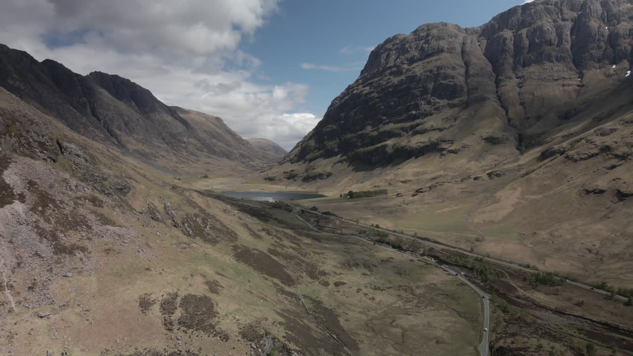 Drone panning right away from Glencoe valley revealing the main road, with snow patches on mountain and dry brown grass