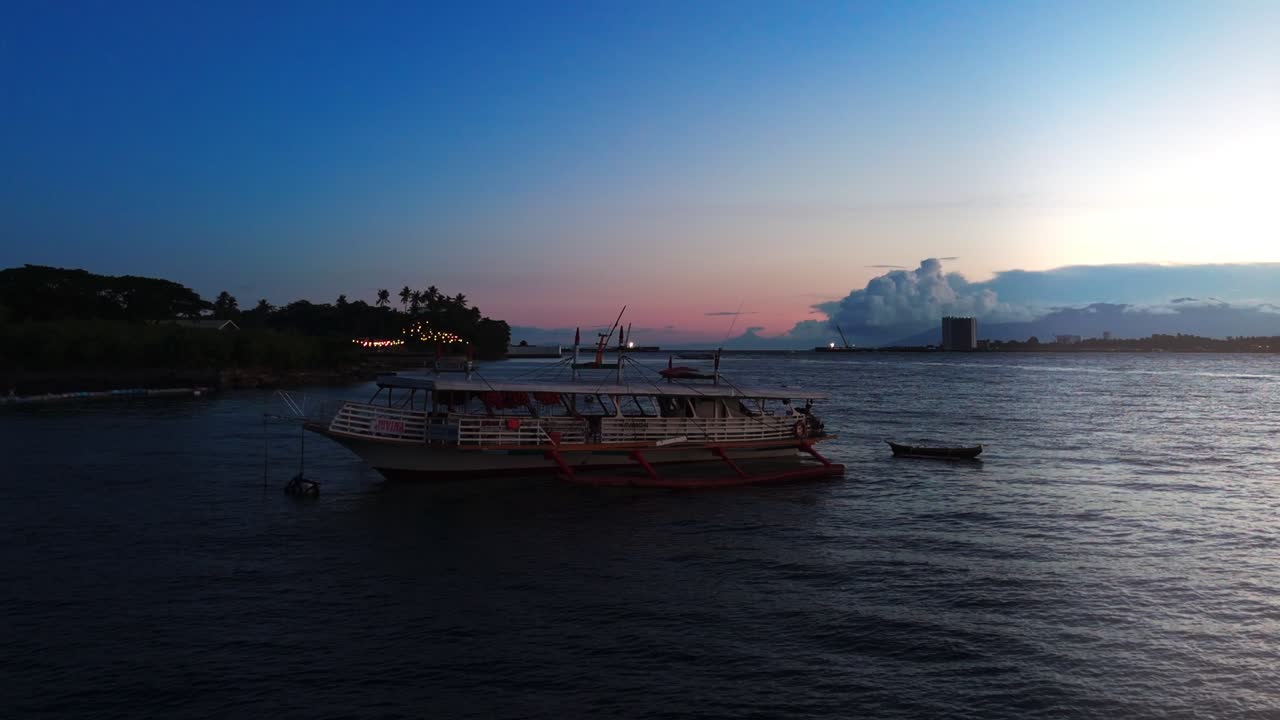Atmospheric aerial footage of a traditional Philippine outrigger boat at dusk with coastal skyline and glowing evening lights. Perfect for travel, culture, and cinematic projects