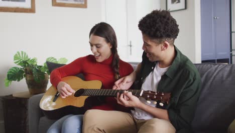 Happy-biracial-couple-sitting-on-sofa-in-living-room-playing-guitar