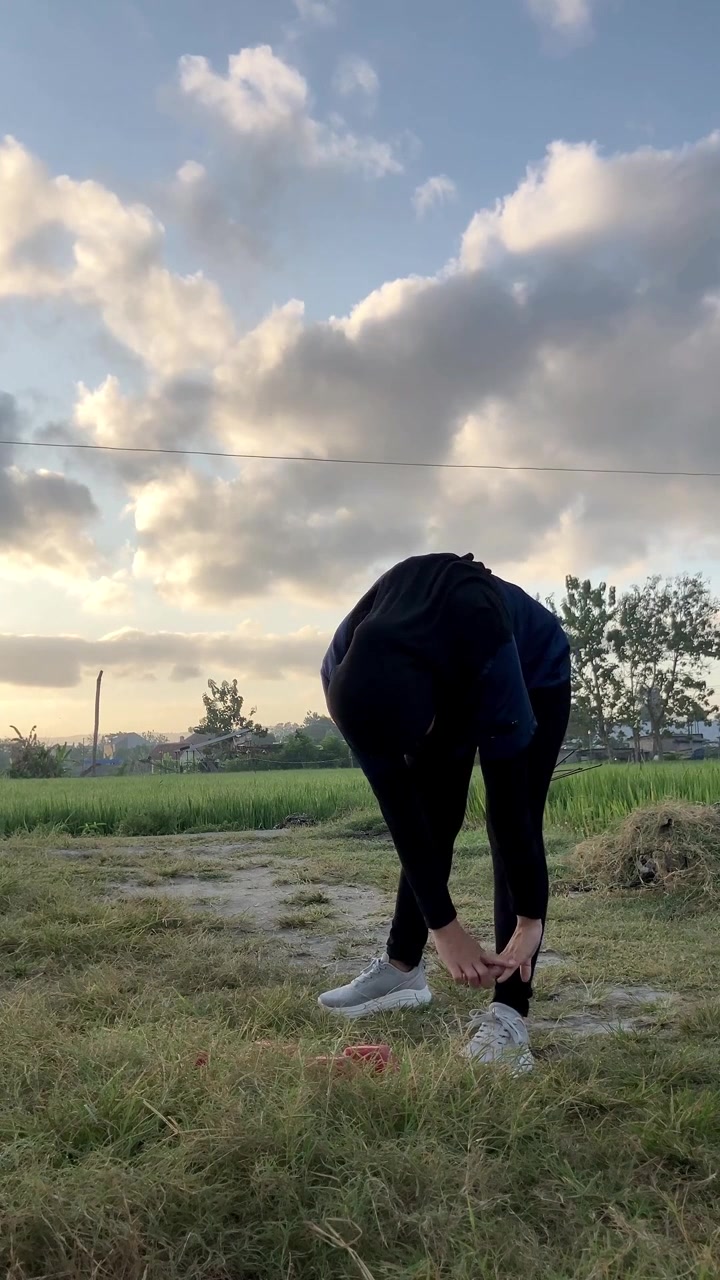 Video verticale al rallentatore di una donna musulmana asiatica con l'hijab che esegue un piegamento in avanti toccandosi le punte dei piedi. Si riscalda all'aperto in un campo verde con un bellissimo cielo al tramonto.