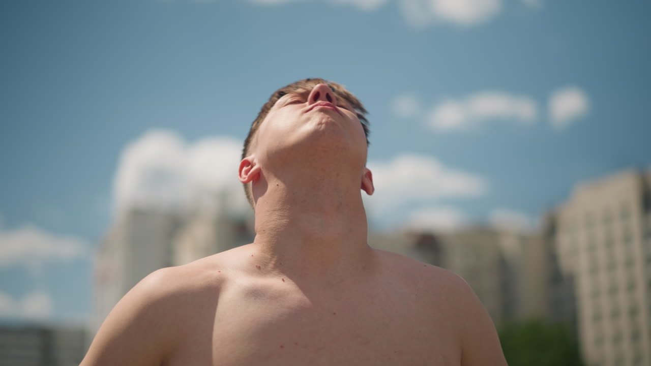 옥상에서 목을 뻗고 있는 20대 남성, 도시 옥상에서 위로 손을 뻗는 밝은 피부의 젊은 남성, 여름날 도시 스카이라인을 배경으로 하늘을 바라보는 활기찬 청년