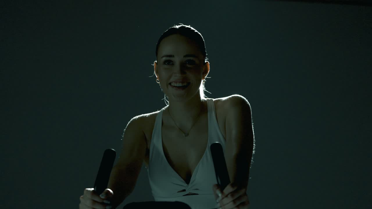 Frontal waist-up shot of a woman smiling gently as she grips the handlebar and begins pedaling. The camera slides slightly from left to right, capturing her motion and energy in soft cinematic light