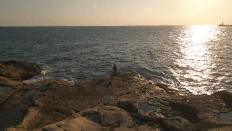 Atemberaubende-Luftaufnahme-Eines-Fischers,-Der-Auf-Flachen-Felsen-Fischt,-Während-Sich-Das-Licht-Der-Untergehenden-Sonne-Auf-Dem-Meer-Bis-Zum-Horizont-Spiegelt,-Malta