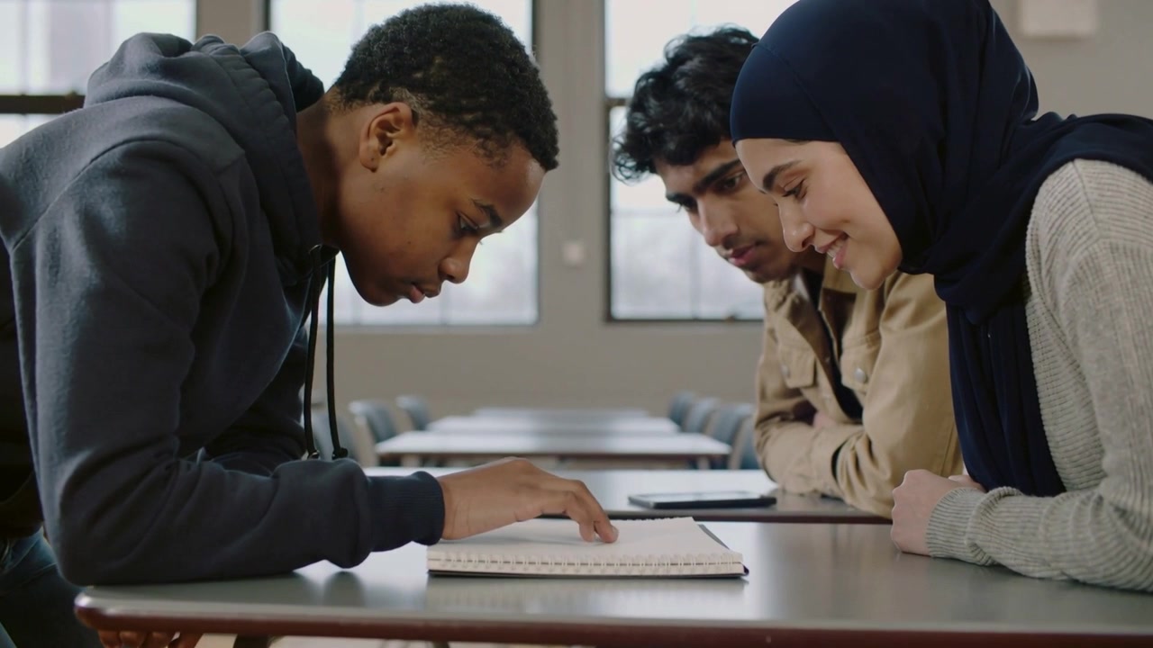 Leaning student in hoodie pointing and tracing notebook at school table, classmates solving problem