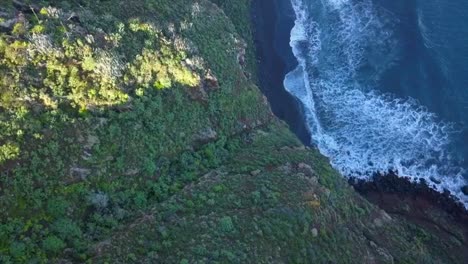 ascenso de drones que revela un acantilado verde cubierto de vegetación tropical en una playa de arena negra volcánica