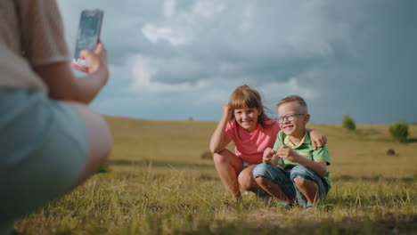 brother and sister squatting outdoors in golden sunlight, smiling as they pose for a photo, sister affectionately holds her brother while their mother captures the heartwarming sibling moment