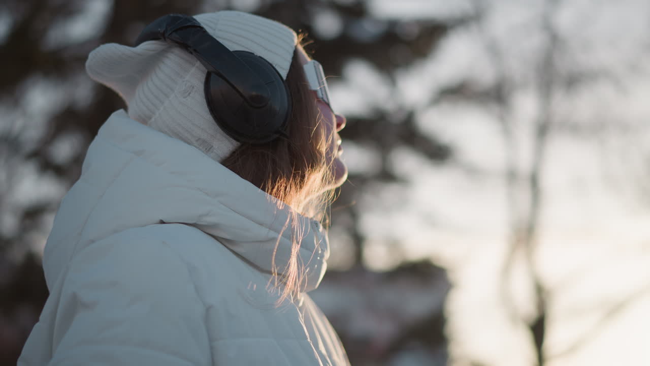 Side view radiant woman smiling in tinted sunglasses under bright winter sky wearing beanie and puffer coat with headphones as breeze tousles hair and light glints on lenses conveying joyful mood