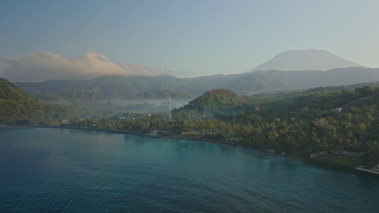 Toàn cảnh bờ biển Bali lúc bình minh với hàng dừa, sóng biển và núi lửa Agung ở phía xa, thể hiện khung cảnh nhiệt đới và phong cảnh thiên nhiên hùng vĩ