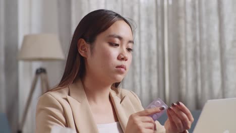 아픈 아시아 사업가 여성이 집에서 거실에서 노트북으로 일하면서 약을 복용하고 있습니다.