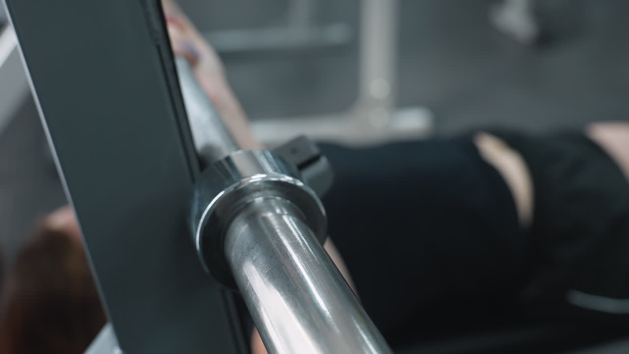 woman rests after lowering barbell onto rack during strength training, close up view of hands gripping metal bar with partial body and background gym equipment slightly visible in blurred focus
