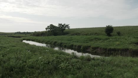 Una-Corriente-De-Agua-Tranquila-Y-Tranquila-En-Medio-De-Un-Campo-De-Pradera-De-Kansas-En-Las-Colinas-De-Pedernal-Rodeadas-De-Hierba-Verde,-Un-Par-De-árboles-Y-Colinas-En-Verano