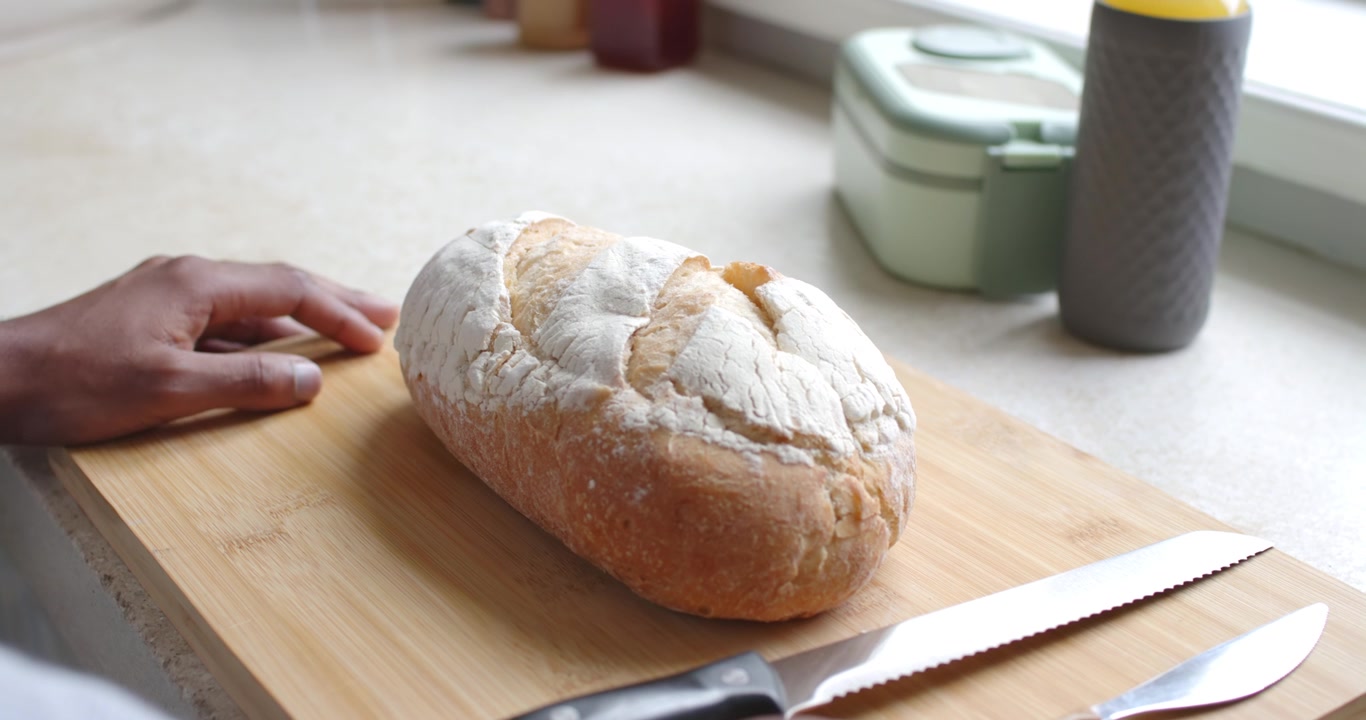 Mani maschili che si muovono da sinistra sul bancone vicino alla finestra, vicino a una pagnotta croccante su un tagliere, per prendere un coltello e preparare.