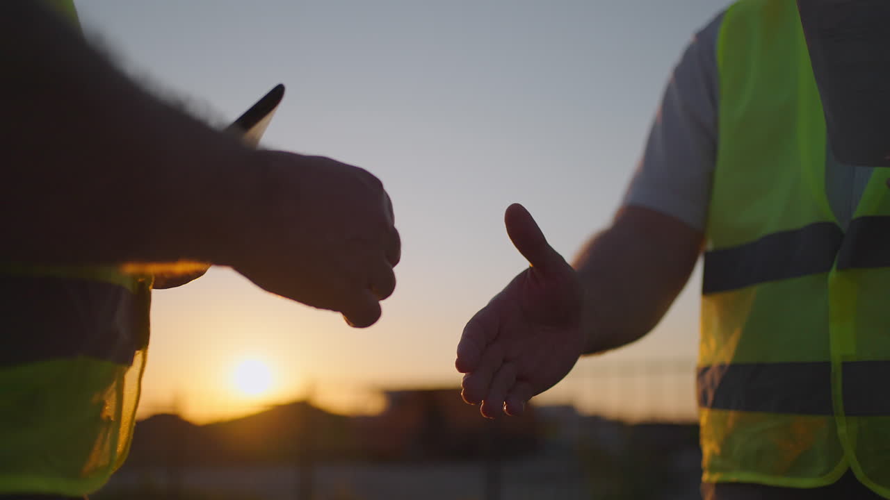 Construction manager and workers shaking hands on construction site. Builder man with a tablet and a man inspector in white helmets shake hands at sunset standing. Symbol of agreement successful work.