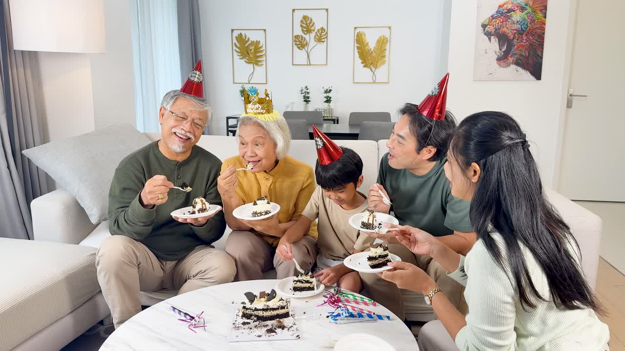 Four generations share birthday cake in a bright living room, smiling and laughing, static camera