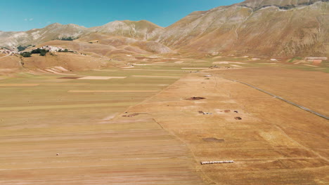 Luftaufnahme-Von-Piana-Grande-Mit-Landschaftsfeldern-Und-Dem-Dorf-Castelluccio-In-Der-Ferne-In-Der-Toskana,-Italien