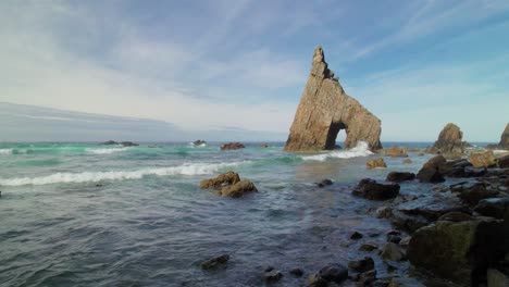 Cinematic-forward-dolly-of-a-wild-pebble-beach-with-sharp-cliffs-and-a-natural-rock-arch-in-the-Cantabrian-Sea-in-Asturias,-Spain