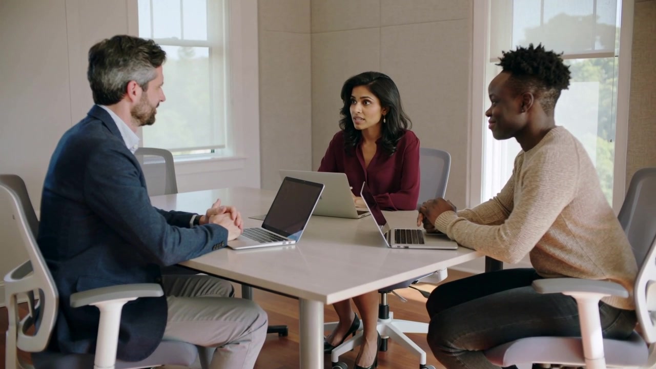 Starting discussion, woman in burgundy blouse presenting plan at office table with three laptops