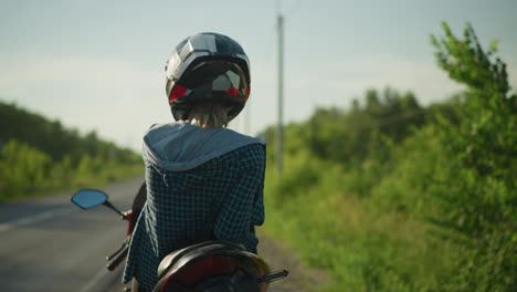 a back view of a biker gazing into the distance on a serene road, surrounded by lush greenery and electric poles, the rider looks contemplative, with the helmet reflecting light