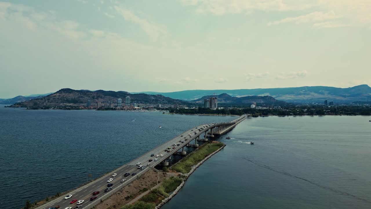 Ripresa aerea in avvicinamento sopra il William R Bennett Bridge, Kelowna, British Columbia, Lago Okanagan, che sale lentamente con panoramica verso il basso mostrando auto, camion, traffico, barche che passano sopra e sotto, città sullo sfondo con le montagne