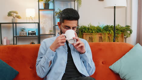 Tired-smiling-Indian-man-drinking-cup-of-warm-coffee-or-herbal-tea-in-the-morning-relaxing-enjoying