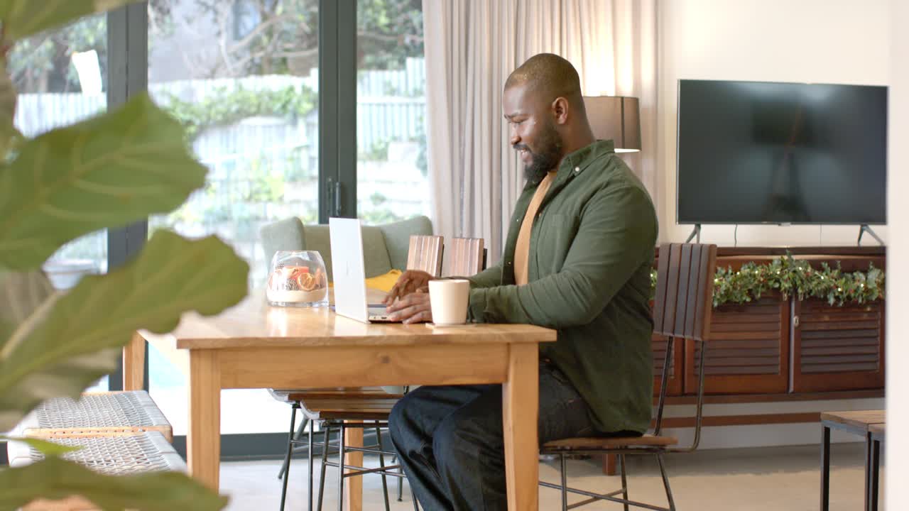 African American mid adult male entering frame sitting at home working on laptop sipping coffee mug