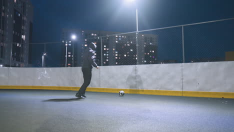 hombre pateando una pelota de fútbol contra la pared bajo la luz de la calle en un campo deportivo urbano iluminado, la pared refleja la silueta, mostrando movimiento atlético dinámico y actividad recreativa urbana por la noche