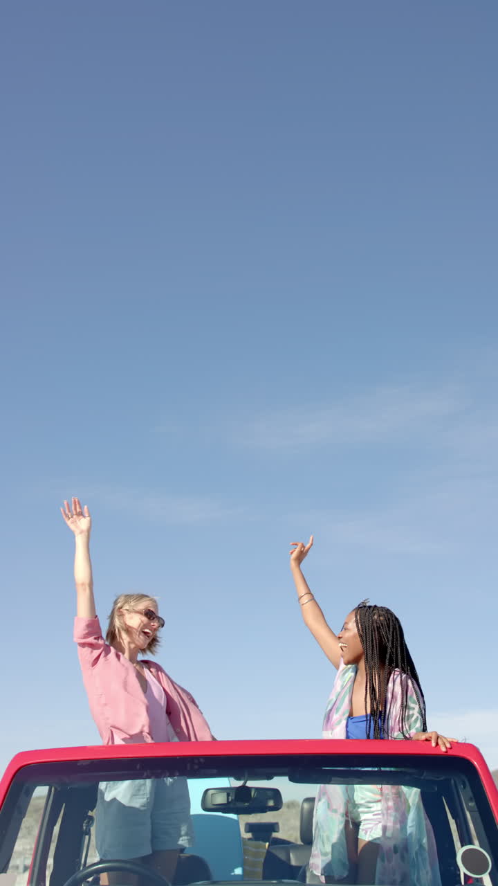 Vertical video: Women enjoying road trip, standing in car with arms raised, smiling