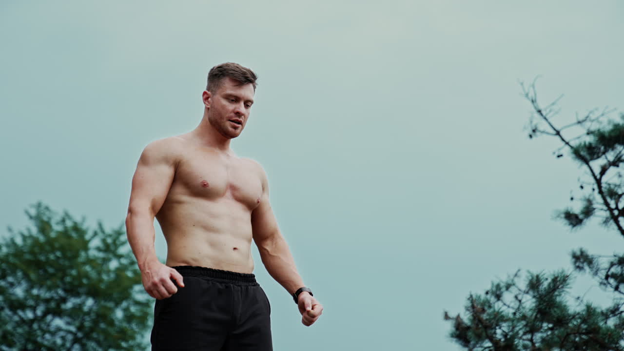 Strong Caucasian man with naked torso stands outdoors. Sportive healthy lifestyle concept. Low angle view.