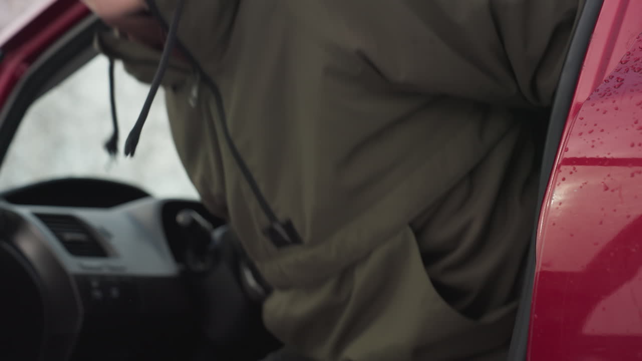 White man wearing winter jacket and sunglasses exits red car onto snowy scene with water droplets on windows and door, capturing crisp cold winter atmosphere and everyday urban movement