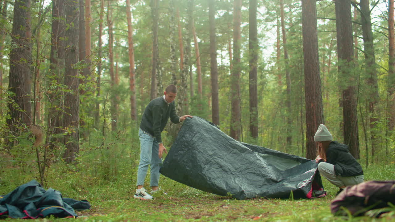 Young couple works together to pack tent after picnic in forest, surrounded by lush greenery and camping bags, leaning in closely as they share affectionate moment during peaceful outdoor departure
