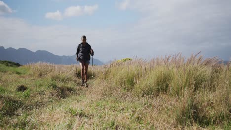 Mixed-race-woman-wearing-backpack-using-nordic-walking-poles-hiking-in-countryside