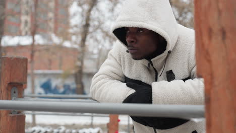 blogger in white hoodie and gloves rests on iron bar outdoors, nodding his head thoughtfully, background features snow-covered ground, trees, and buildings in the distance on a cold winter day