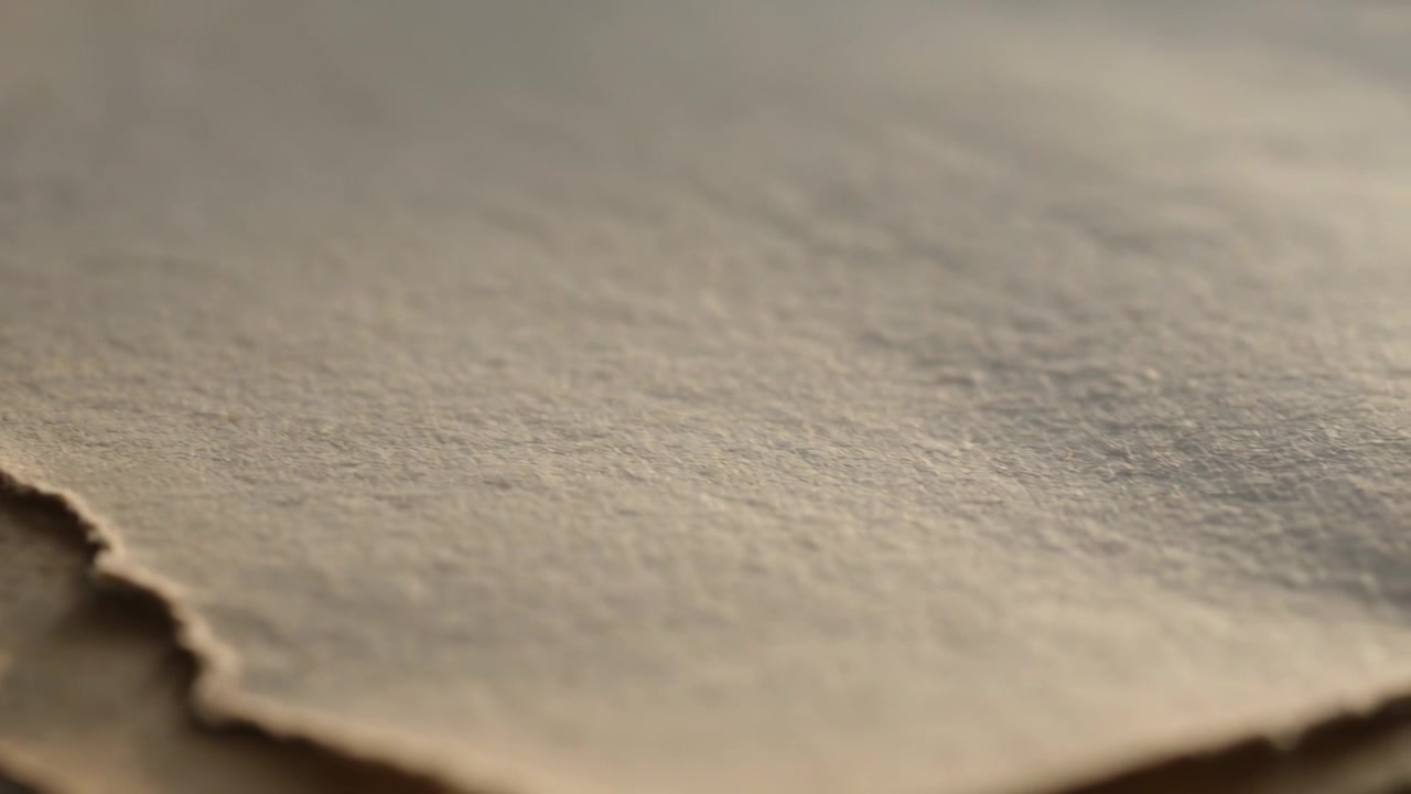 Shifting camera bringing torn deckled paper stack into focus on table, warm light revealing texture