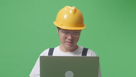 close up of asian man worker wearing goggles and safety helmet smiling and pointing to a laptop in his hand while standing in the green screen background studio