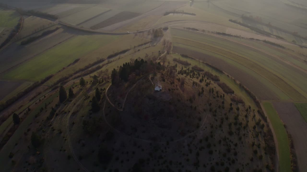 Vista aérea de una iglesia en una colina con vistas a campos y un valle neblinoso, Alemania.