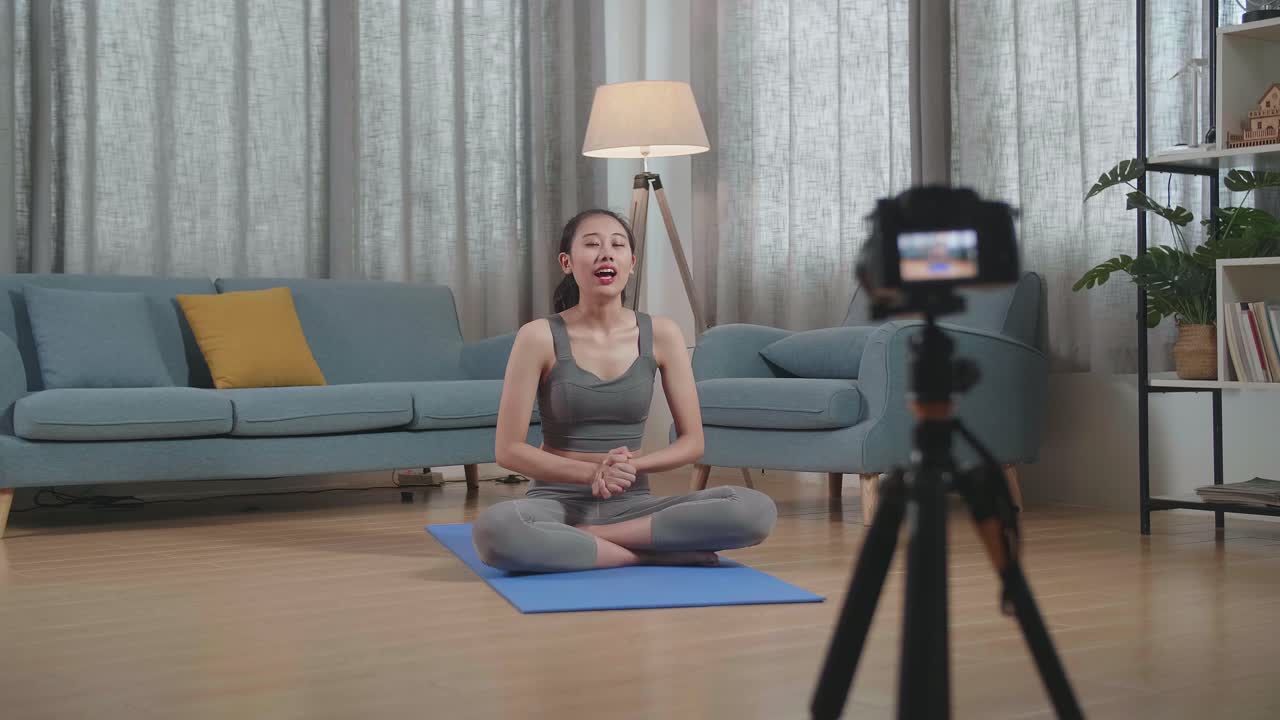 vista completa de joven entrenadora asiática mujer en ropa deportiva agitando la mano y hablando a la cámara mientras graba ejercicio de enseñanza en casa