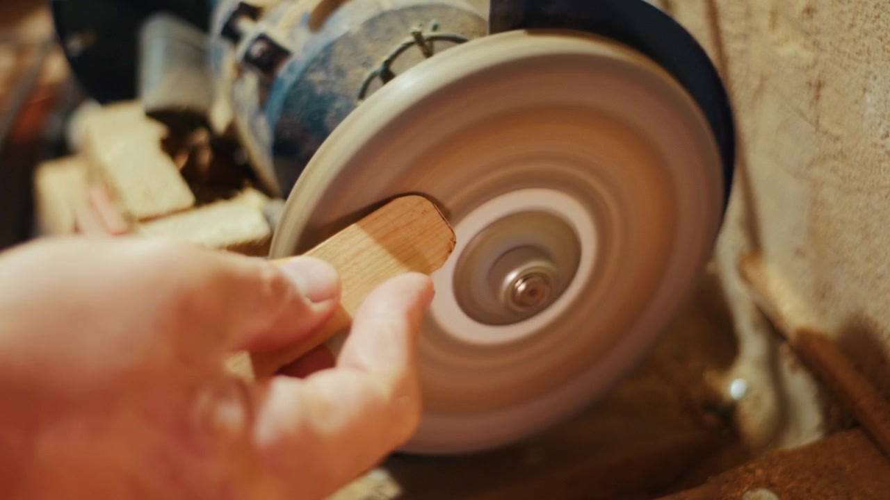 Close-up of a craftsman using a bench grinder to shape and smooth a small piece of wood in a workshop. Authentic DIY woodworking footage for craft, carpentry and maker projects